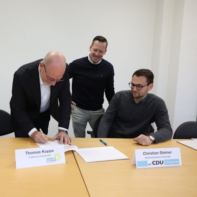 Eine Frau und drei Männer an einem Tisch, einer unterschreibt ein Papier.