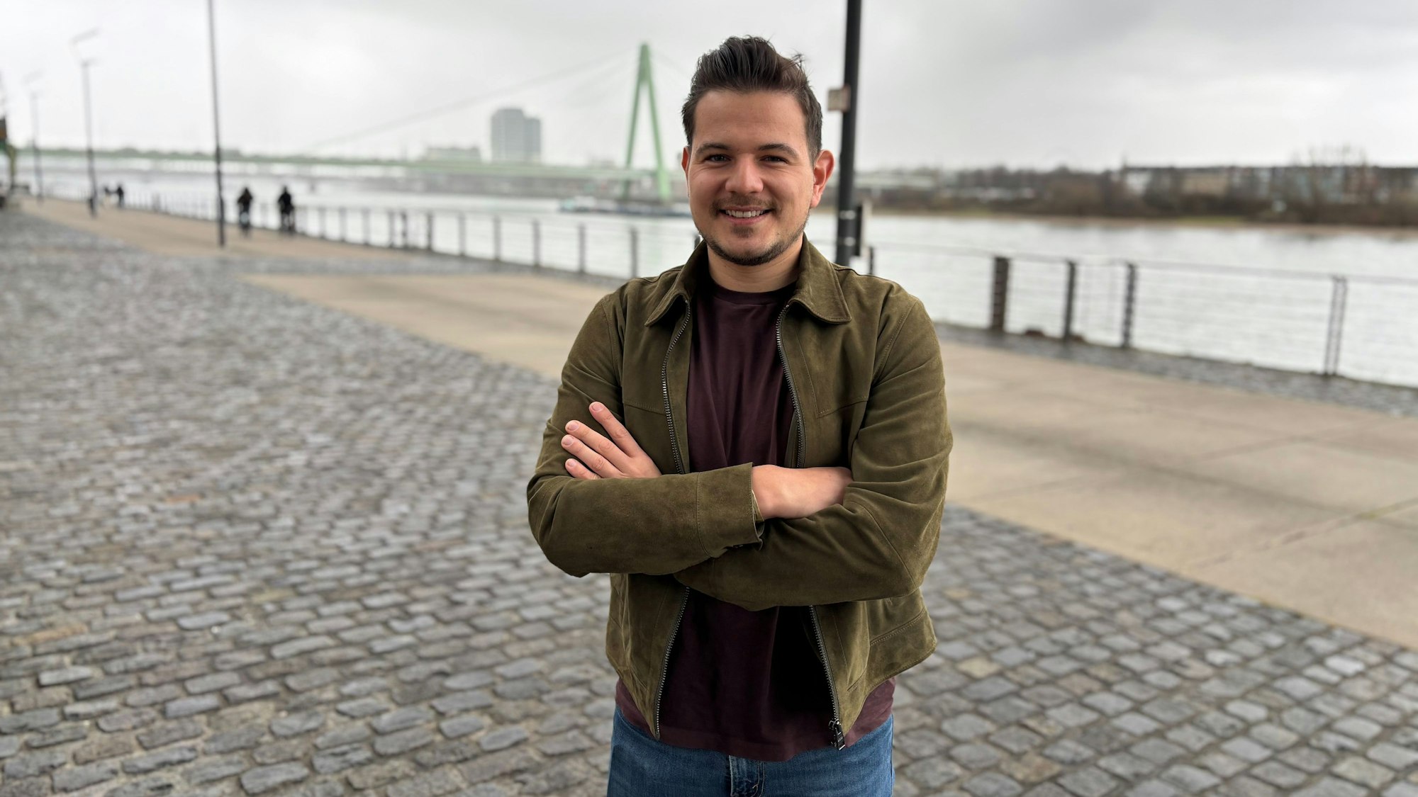 Luis Teichmann steht lächelnd mit verschränkten Armen am Rheinufer.