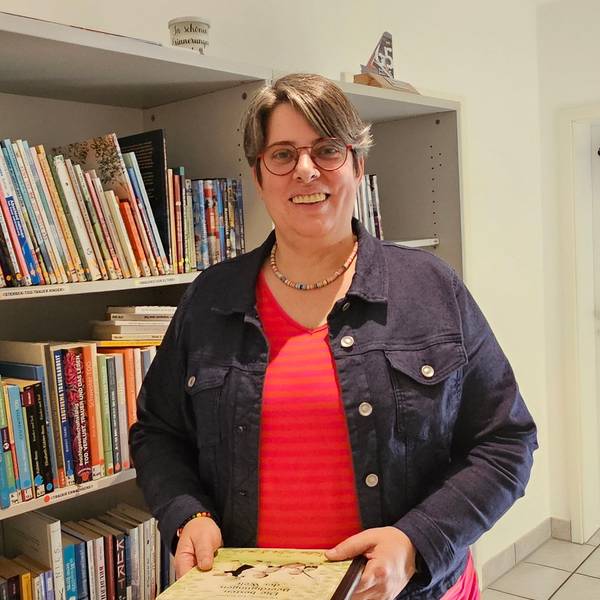 Eine Frau steht in einem Raum und hält ein Buch in der Hand, hinter ihr ist ein Bücherregal.