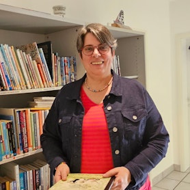 Eine Frau steht in einem Raum und hält ein Buch in der Hand, hinter ihr ist ein Bücherregal.