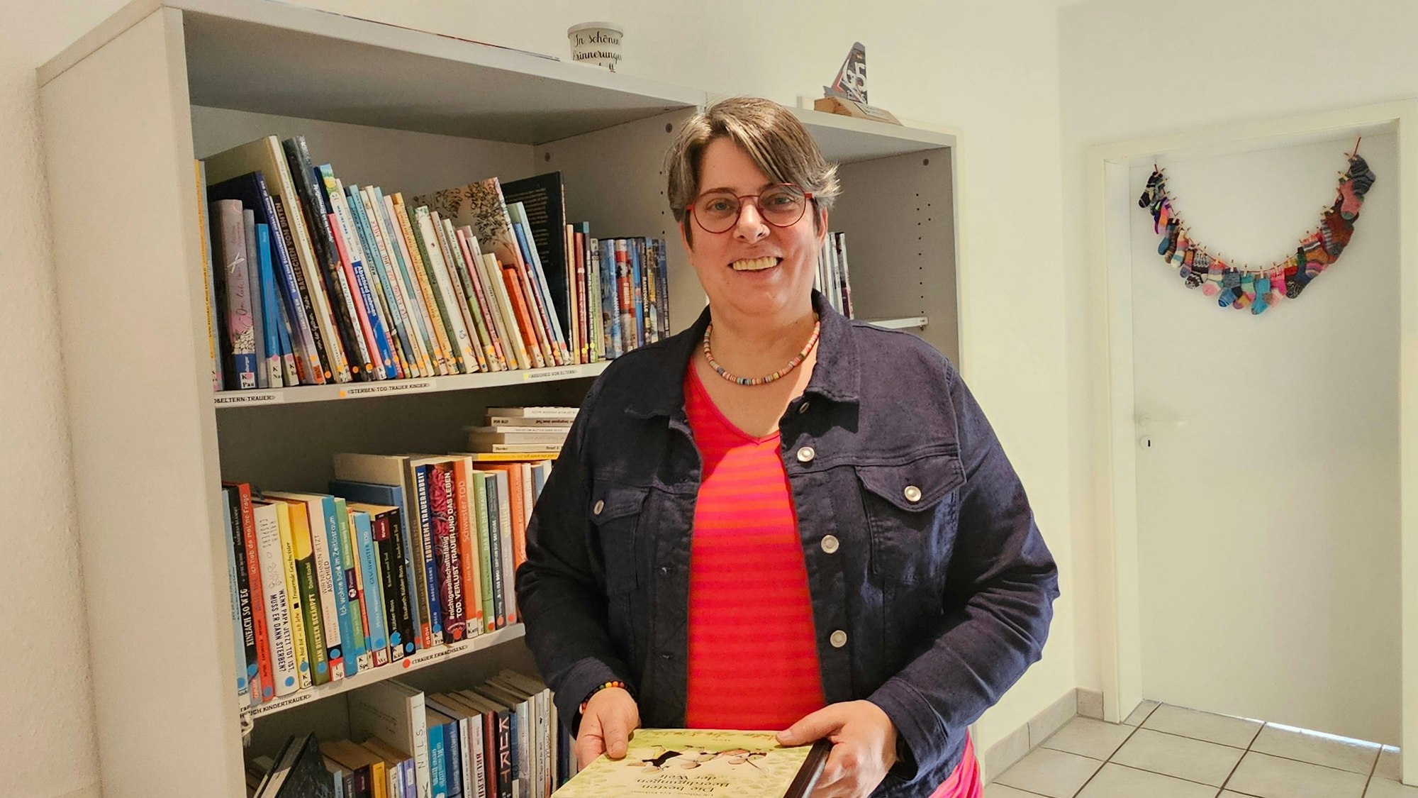 Eine Frau steht in einem Raum und hält ein Buch in der Hand, hinter ihr ist ein Bücherregal.