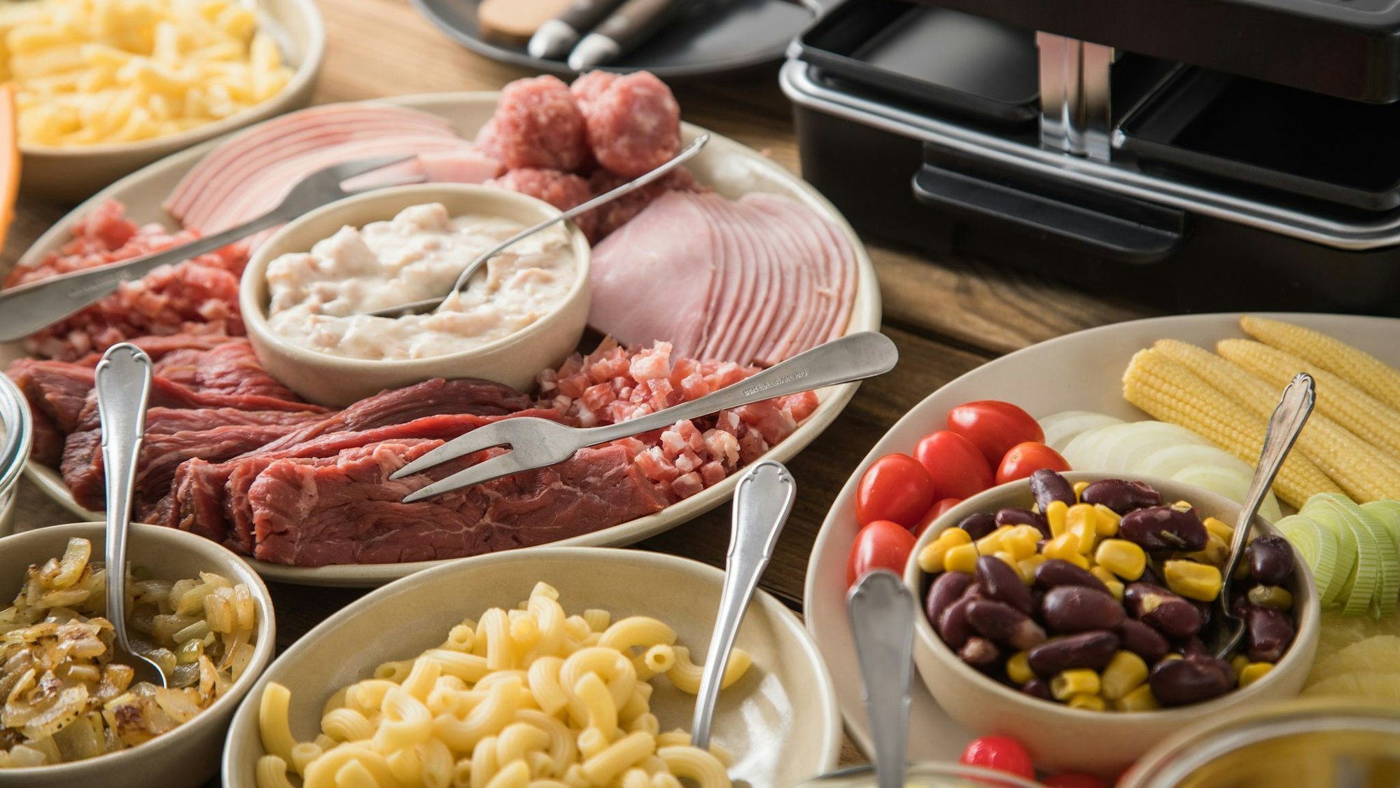 Viele Schälchen und Teller mit Zutaten für Raclette stehen auf einem Tisch.