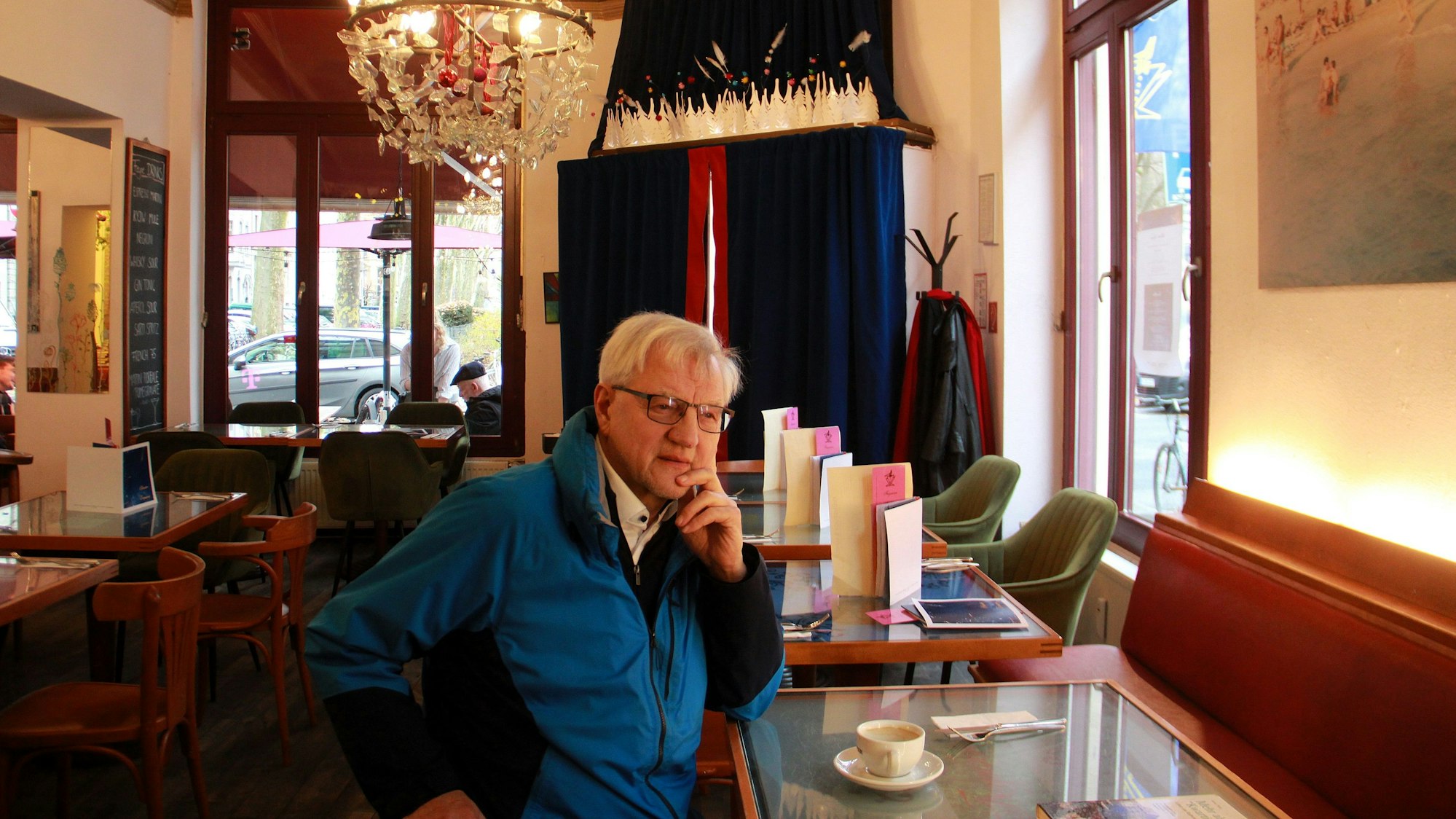 Ein Mann in blauem Anorak sitzt an einem Hochtisch in einem Café. An der Decke hängt ein großer Kristall-Kerzenleuchter.