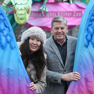 Eine Frau mit Fellmütze und ein Mann im grauen Anzug schauen zwischen zwei großen bunten Flügeln hindurch in die Kamera.