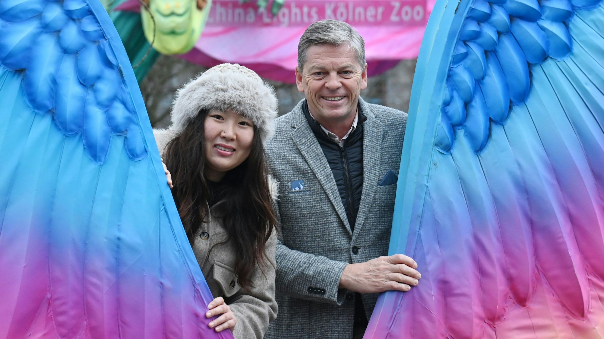 Eine Frau mit Fellmütze und ein Mann im grauen Anzug schauen zwischen zwei großen bunten Flügeln hindurch in die Kamera.