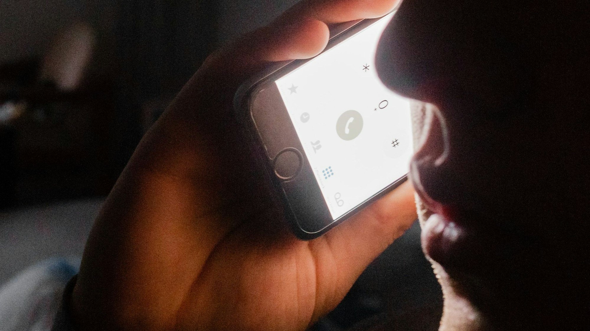 Das Symbolbild zeigt einen Gesichtsausschnitt im Halbdunkel, dazu eine Hand, die ein Telefon hält.