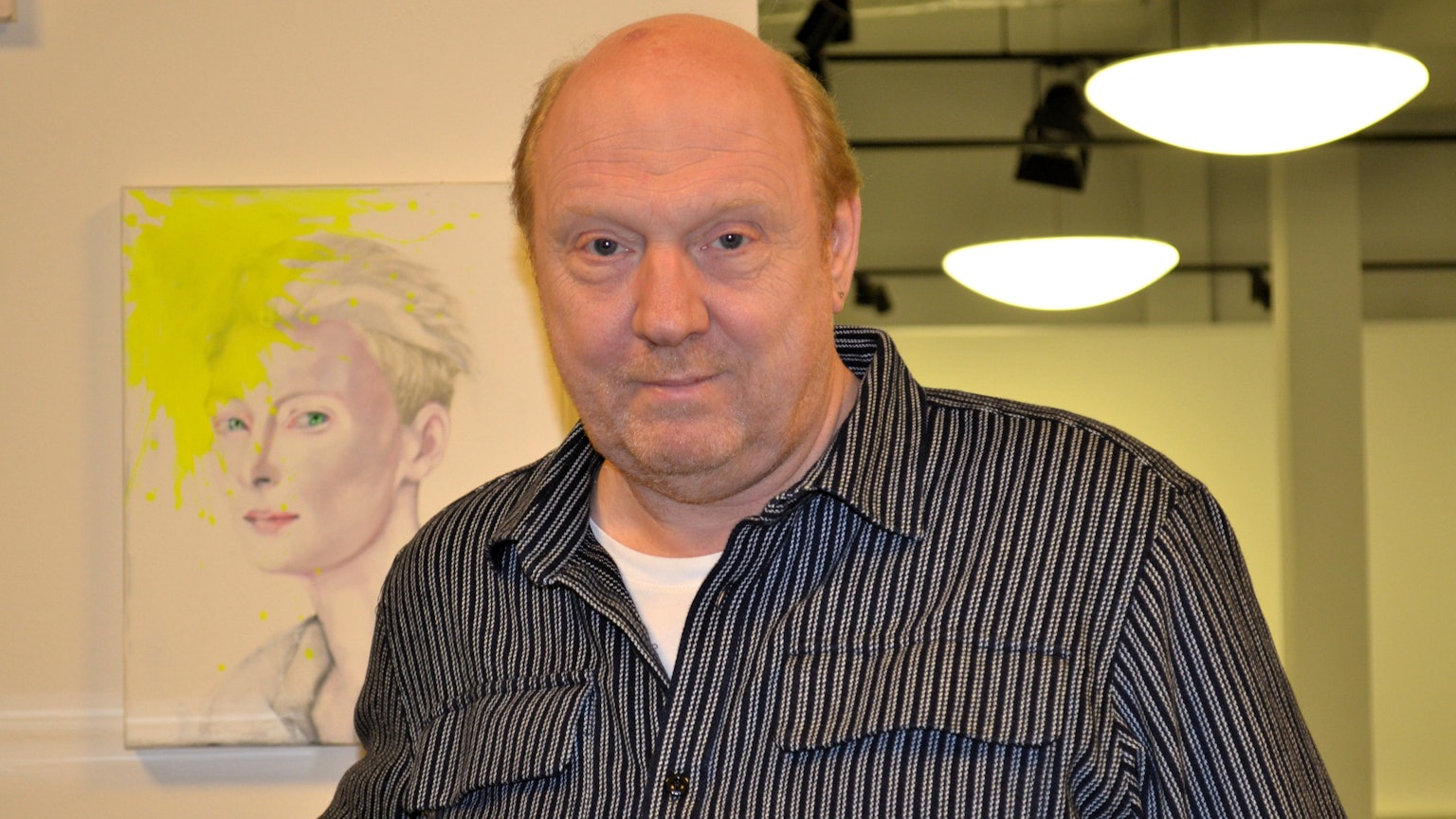 Ein Mann im schwarz-grau gestreiften Hemd vor einer Wand mit einem Gemälde.