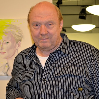 Ein Mann im schwarz-grau gestreiften Hemd vor einer Wand mit einem Gemälde.