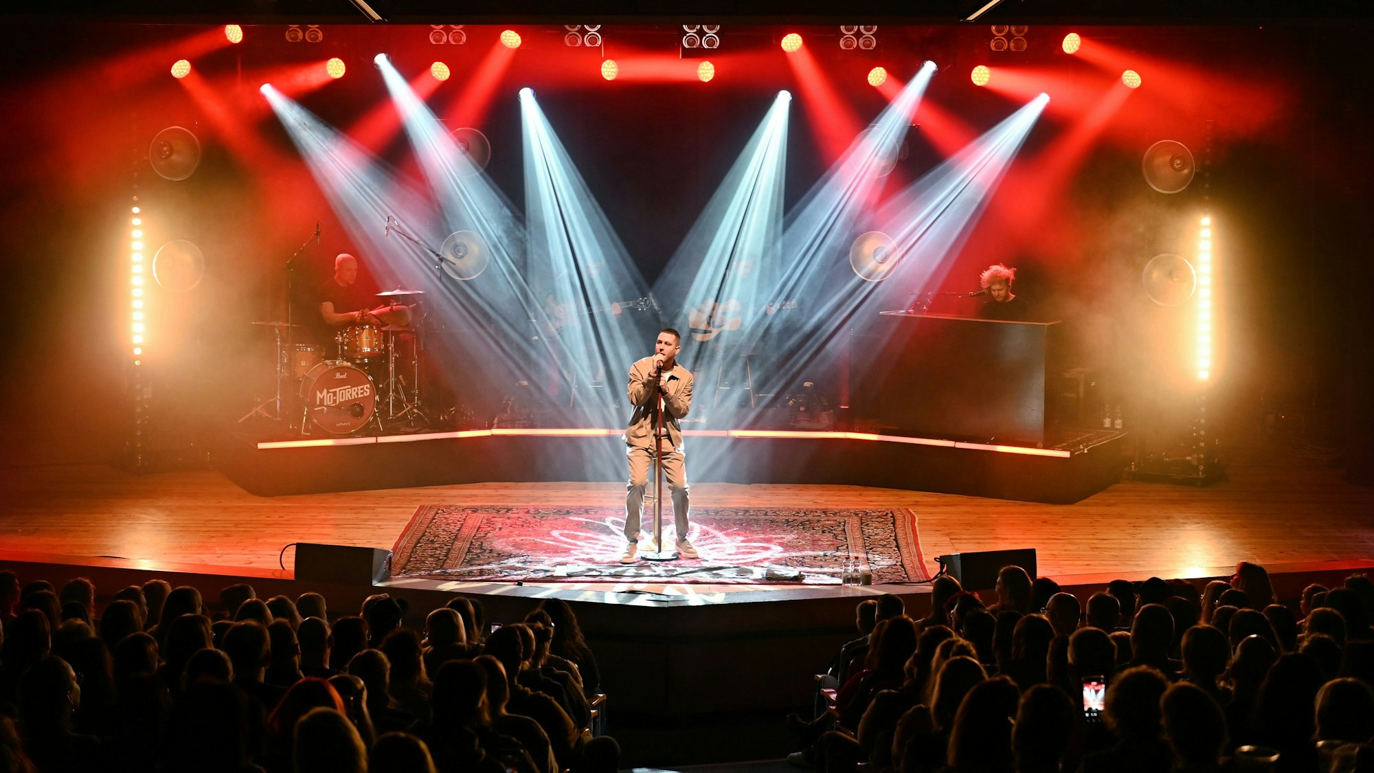 Der Musiker Mo-Torres steht von mehreren Scheinwerfern angeleuchtet auf einer Bühne. Das Publikum vor ihm ist in Umrissen zu sehen.