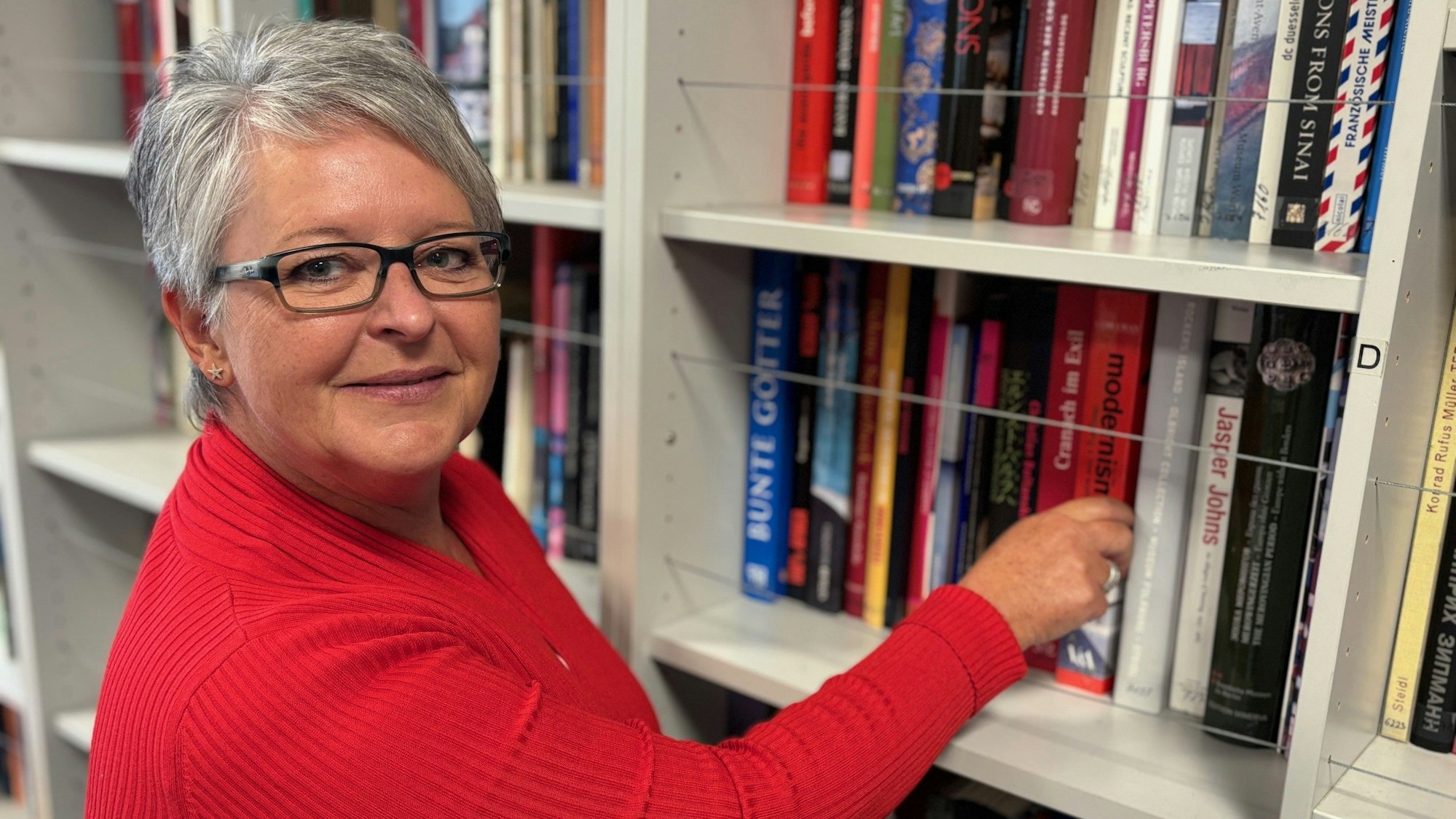 Auf dem Bild ist eine Frau in einem roten Oberteil vor einem Bücherregal zu sehen.