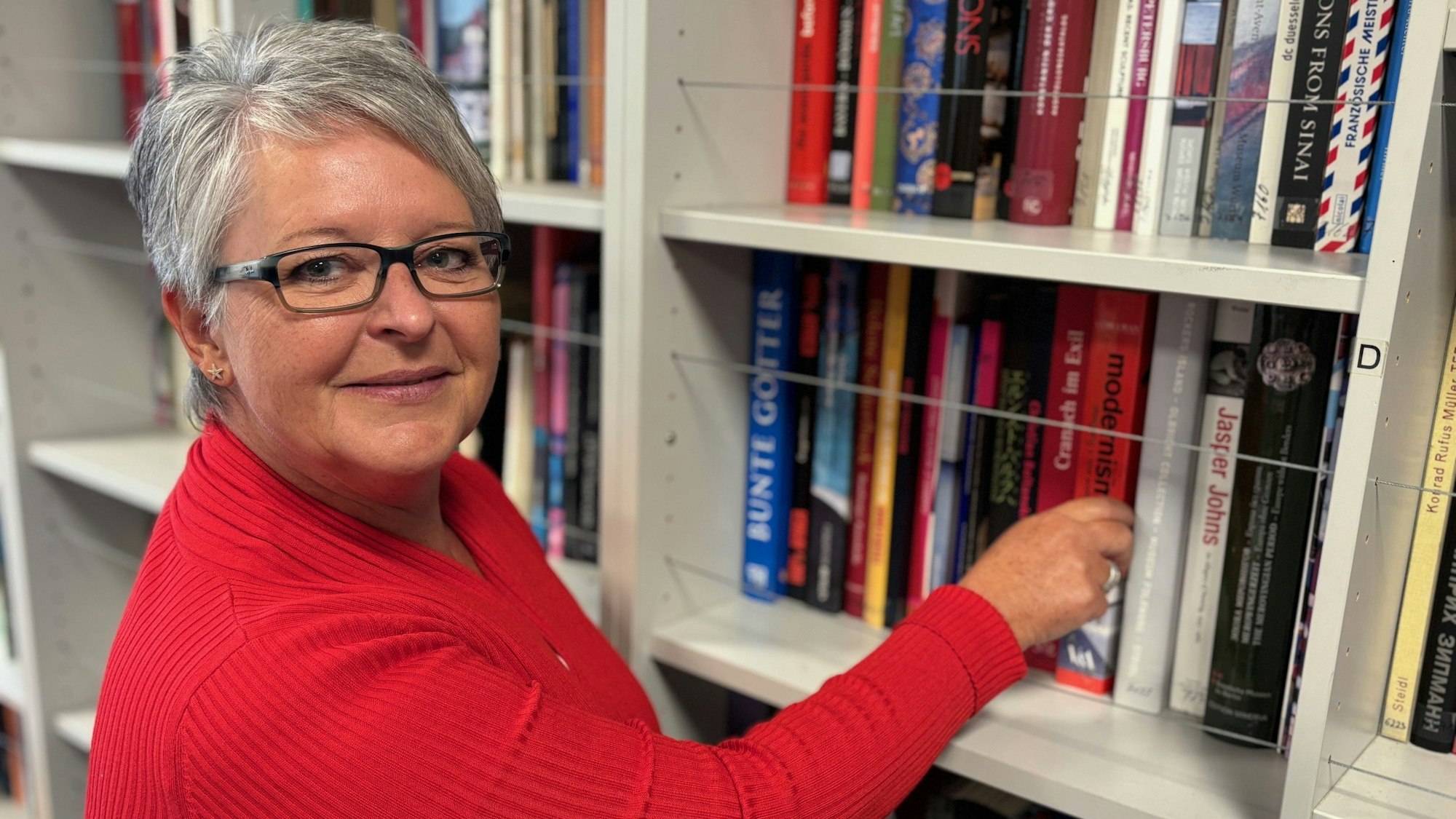 Auf dem Bild ist eine Frau in einem roten Oberteil vor einem Bücherregal zu sehen.