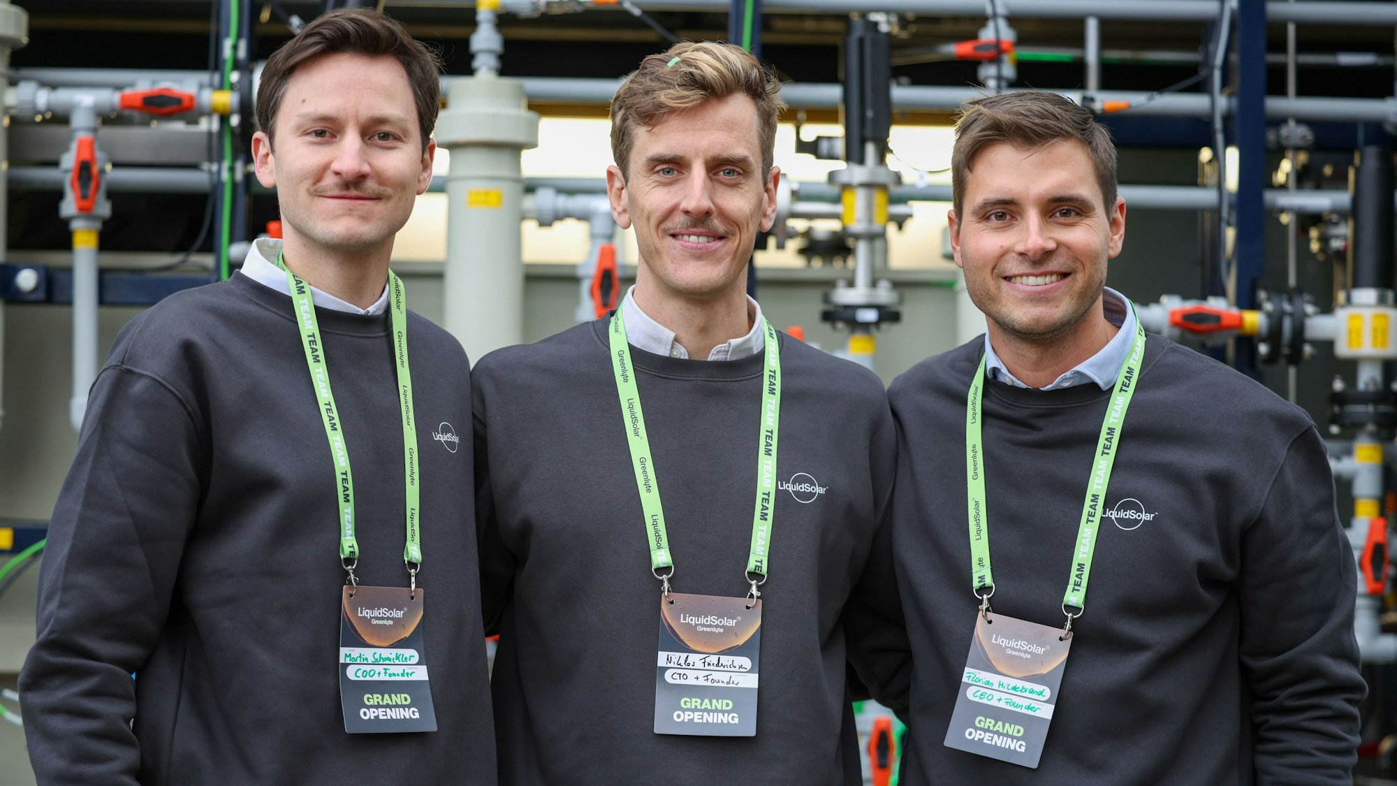 Drei Männer in dunklen Pullovern stehen eng aneinander und lächeln in die Kamera.