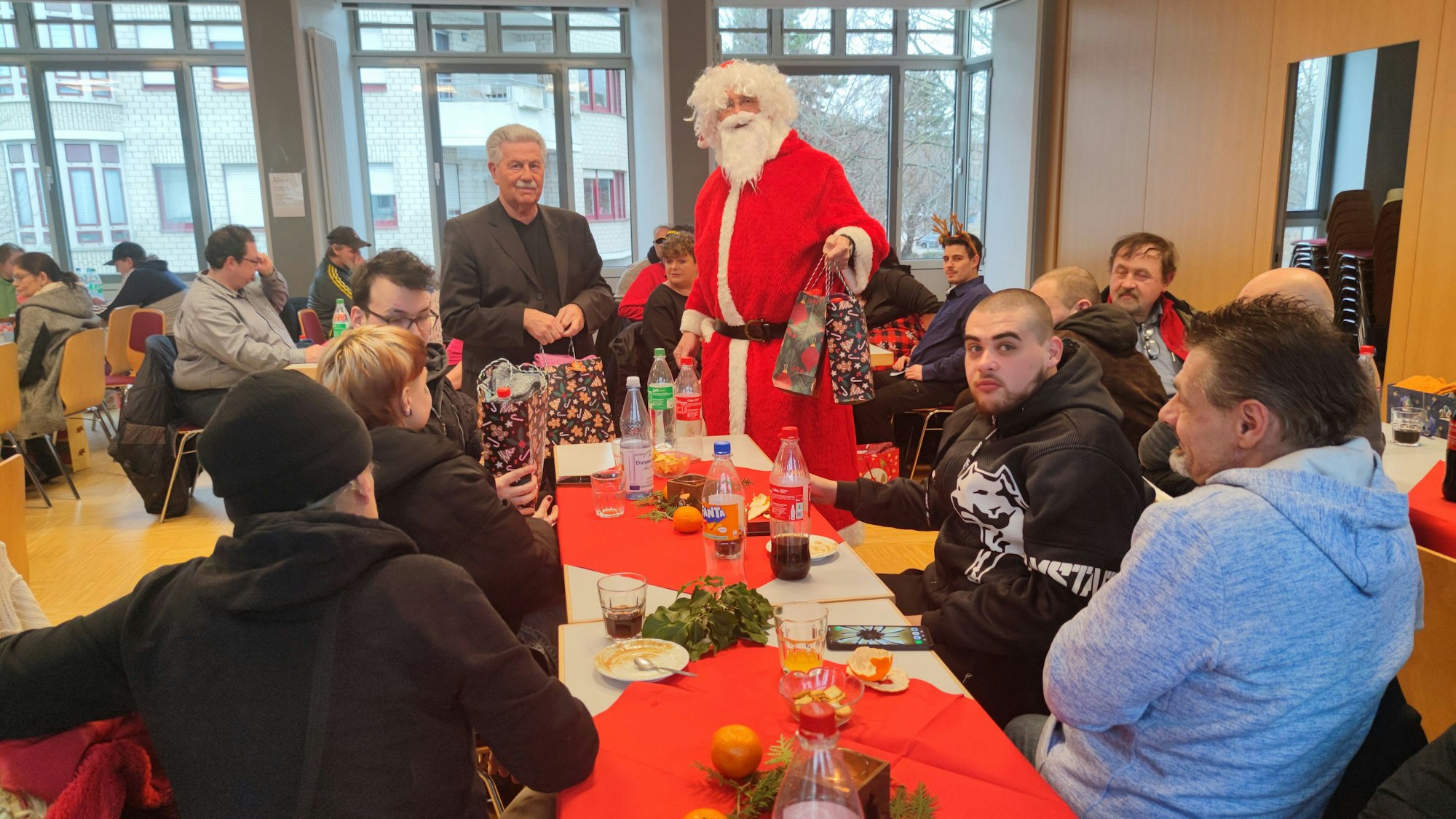 Gäste der Weihnachtsfeier der Wohnungslosenhilfe sitzen an geschmückten Tischen zusammen. Ein Mann im Nikolauskostüm und Hubert Hamacher verteilen die Geschenktüten.