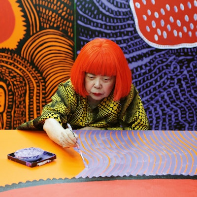 Yayoi Kusama sitzt an einem Tisch und bemalt eine Leinwand.