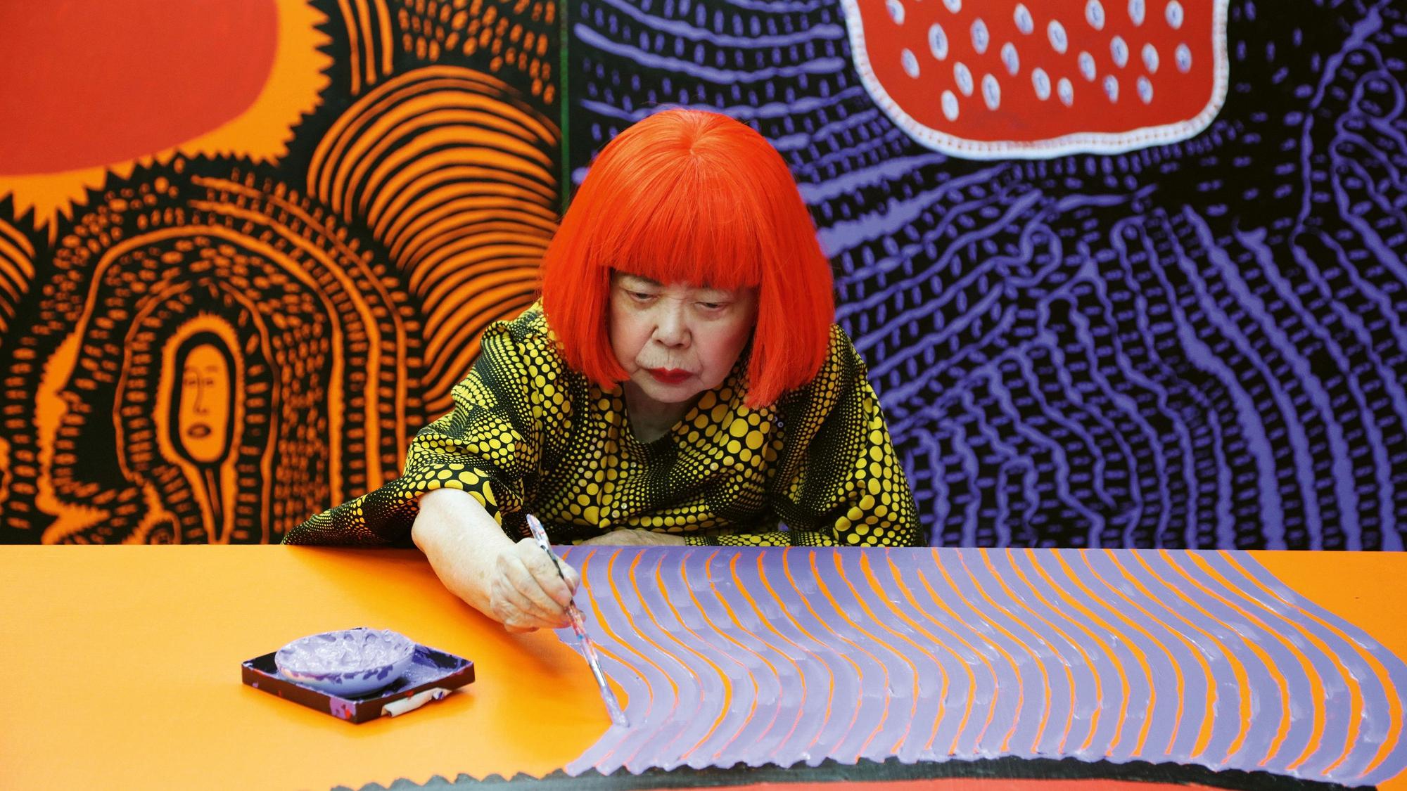 Yayoi Kusama sitzt an einem Tisch und bemalt eine Leinwand.