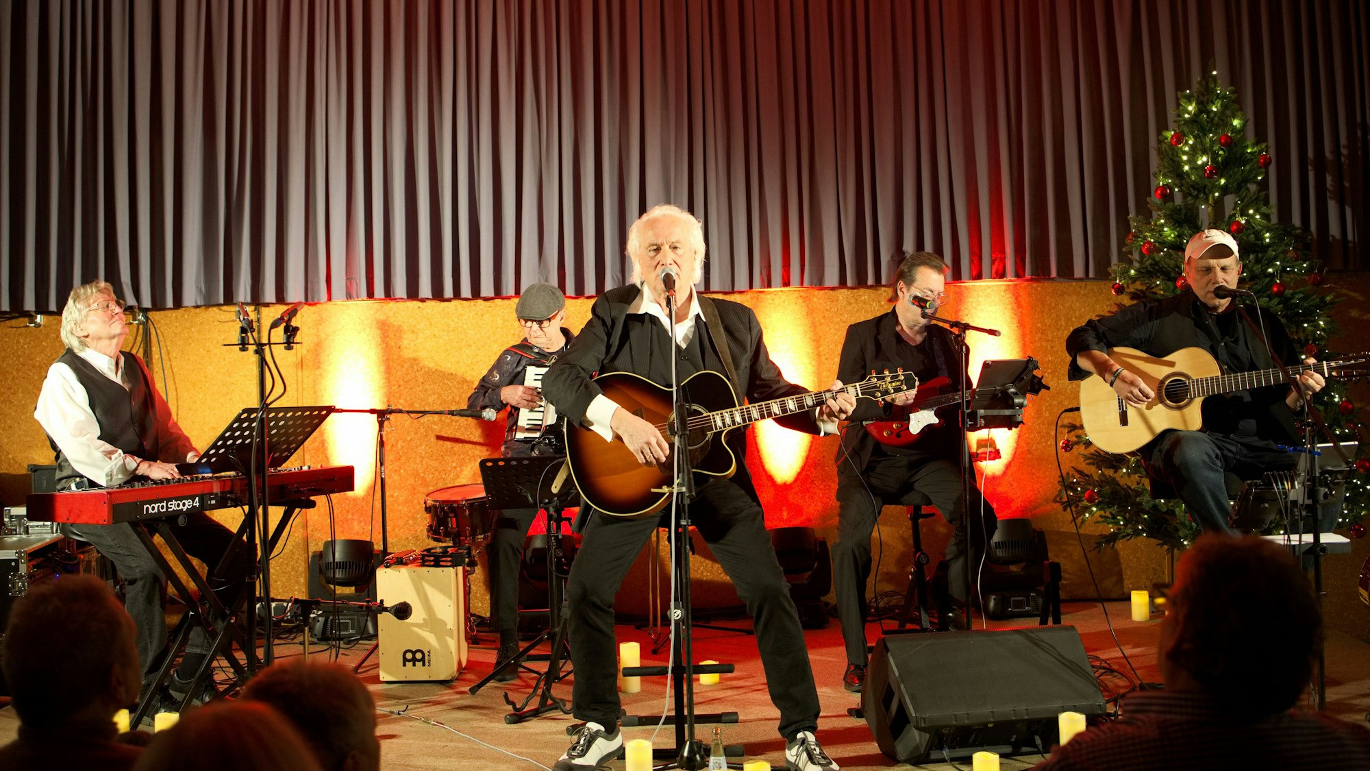 Hannes Schöner steht in der Mitte einer Bühne und singt in ein Mikrofon. Neben und hinter ihm sind weitere Musiker, rechts im Hintergrund ein Weihnachtsbaum.