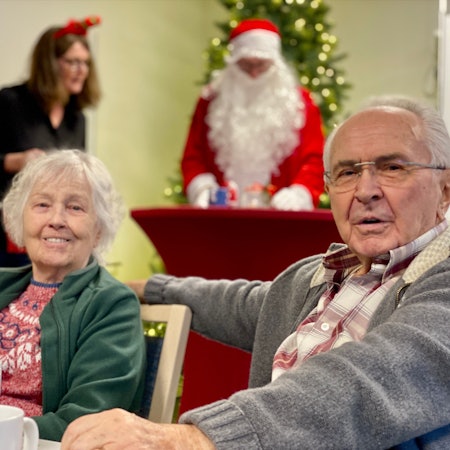 Das Bild zeigt zwei Senioren im Vordergrund. Im Hintergrund ist ein Weihnachtsmann zu sehen.