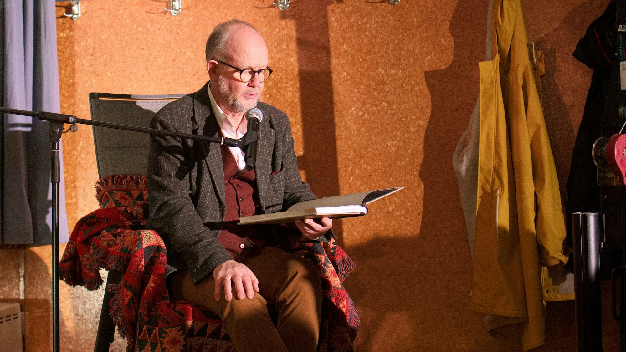 Der Autor Ralf Kramp sitzt in einem Sessel und liest aus einem Buch vor.