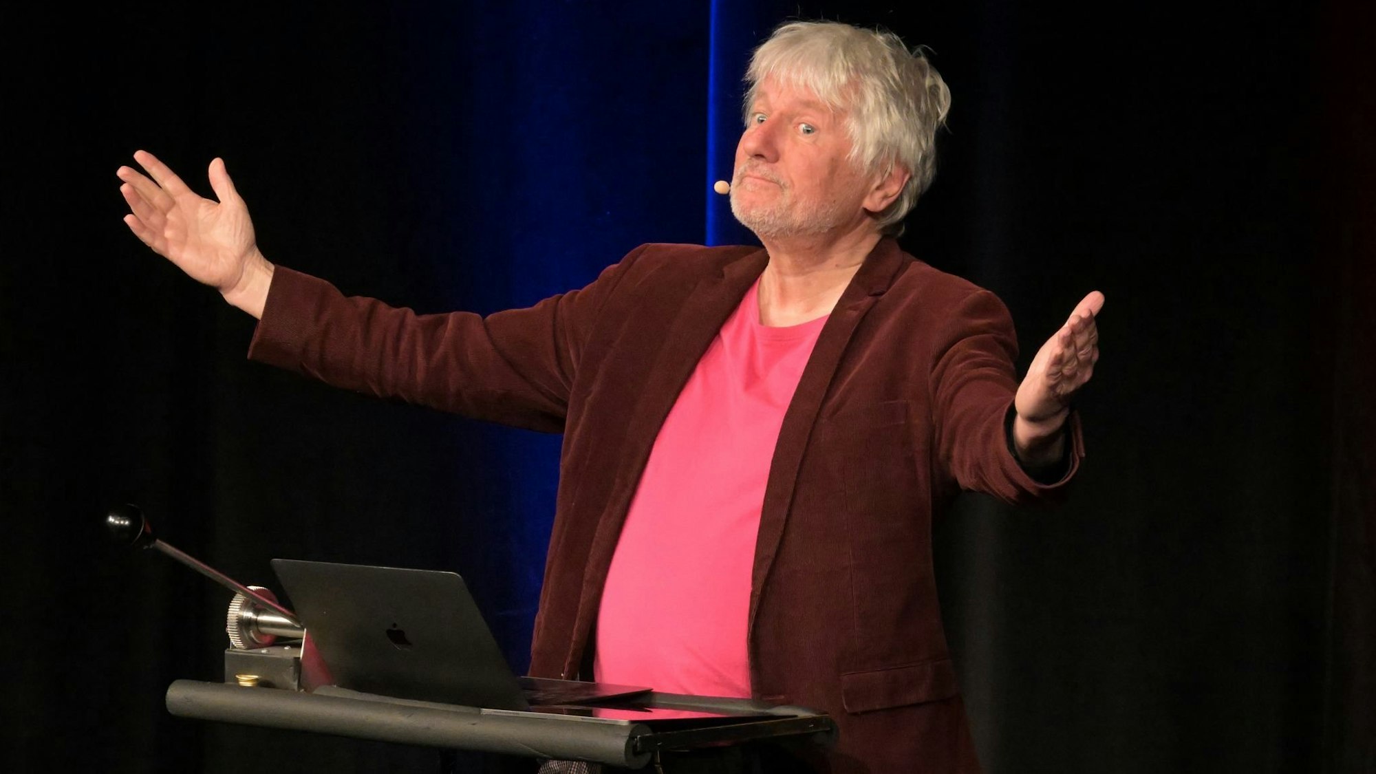 Der Kabarettist Jürgen Becker steht auf einer Bühne und spricht.