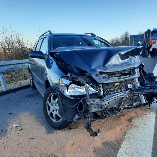 Ein blaues Auto mit demolierter Frontpartie, im Hintergrund ein Einsatzfahrzeug der Feuerwehr.