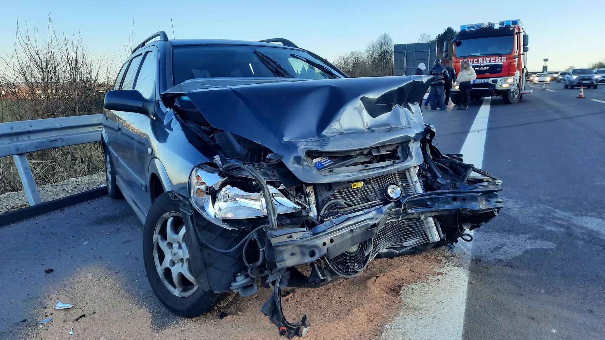 Ein blaues Auto mit demolierter Frontpartie, im Hintergrund ein Einsatzfahrzeug der Feuerwehr.