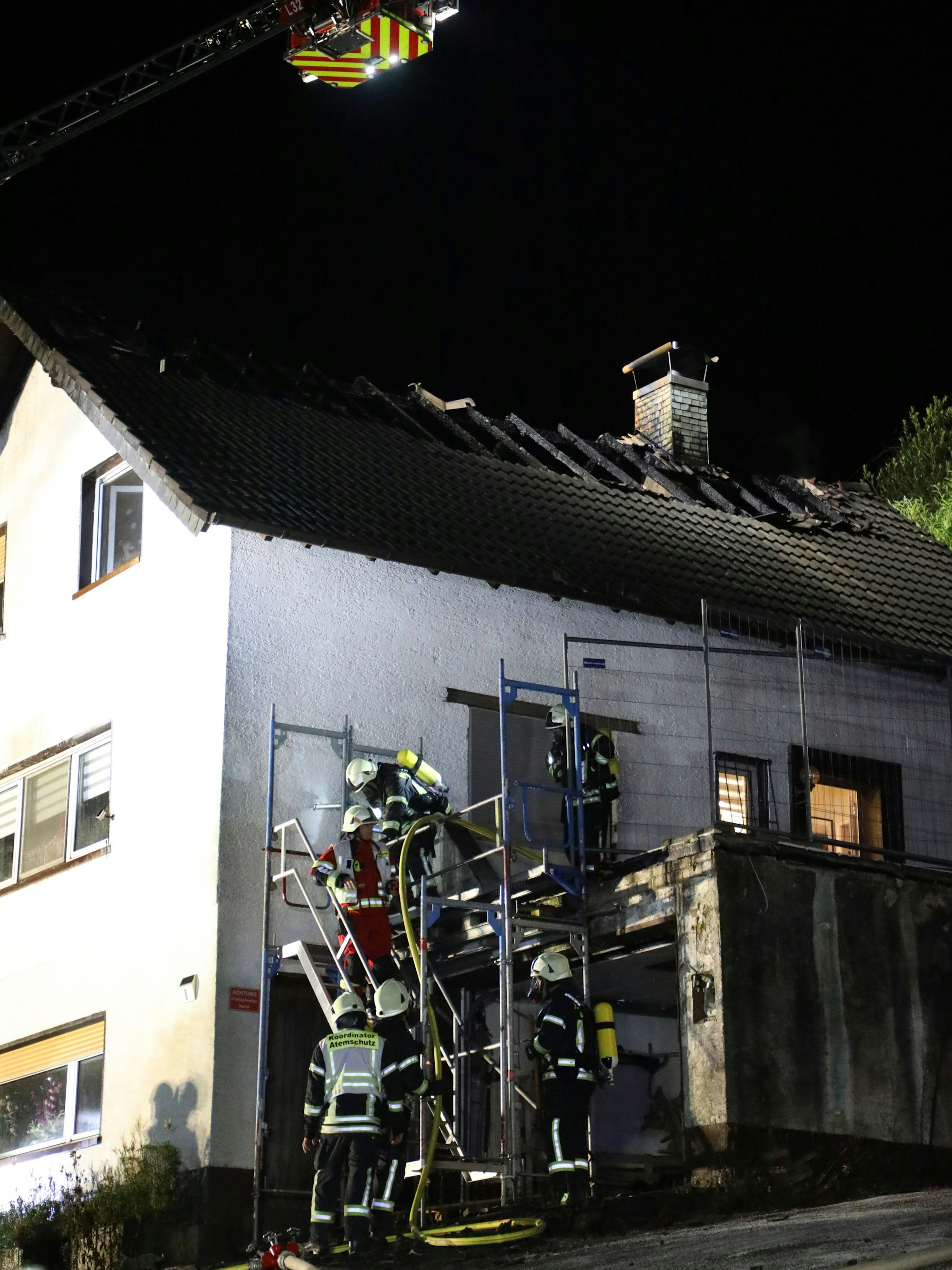 Feuerwehrleute steigen über ein Gerüst zu einem Haus hinauf. Das Dach des Hauses ist abgebrannt.