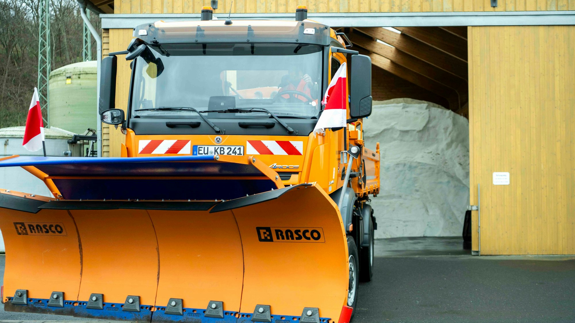 Ein orangefarbenes Schneeräumfahrzeug steht abfahrbereit vor der Halle, in der ein Berg Streusalz zu sehen ist.