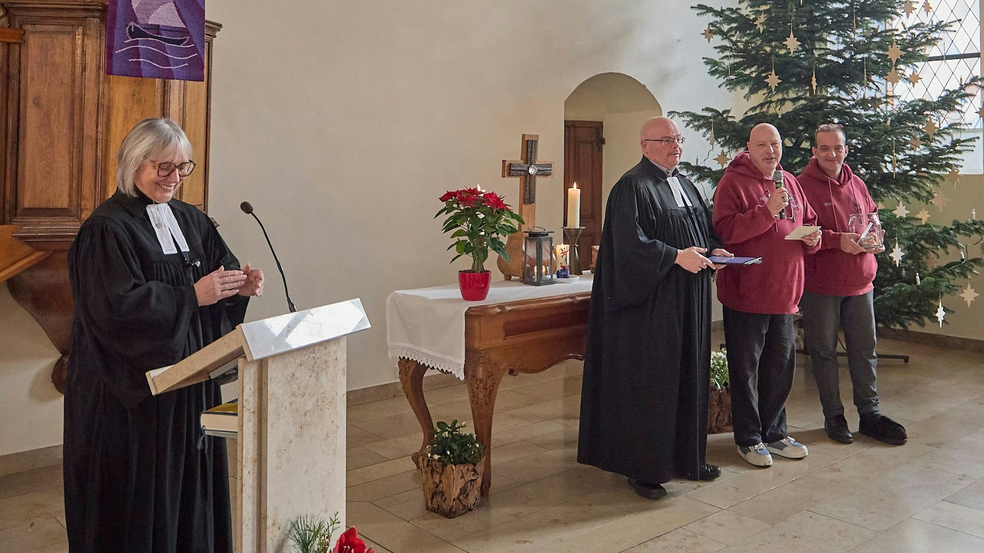 Im Altarraum der Kirche steht Pfarrerin Gaby Leufgen am Redepult, vor dem Altar stehen Oliver Joswig, Peter Schäfer und Heiko Schäfer. Im Hintergrund steht ein großer geschmückter Weihnachtsbaum.