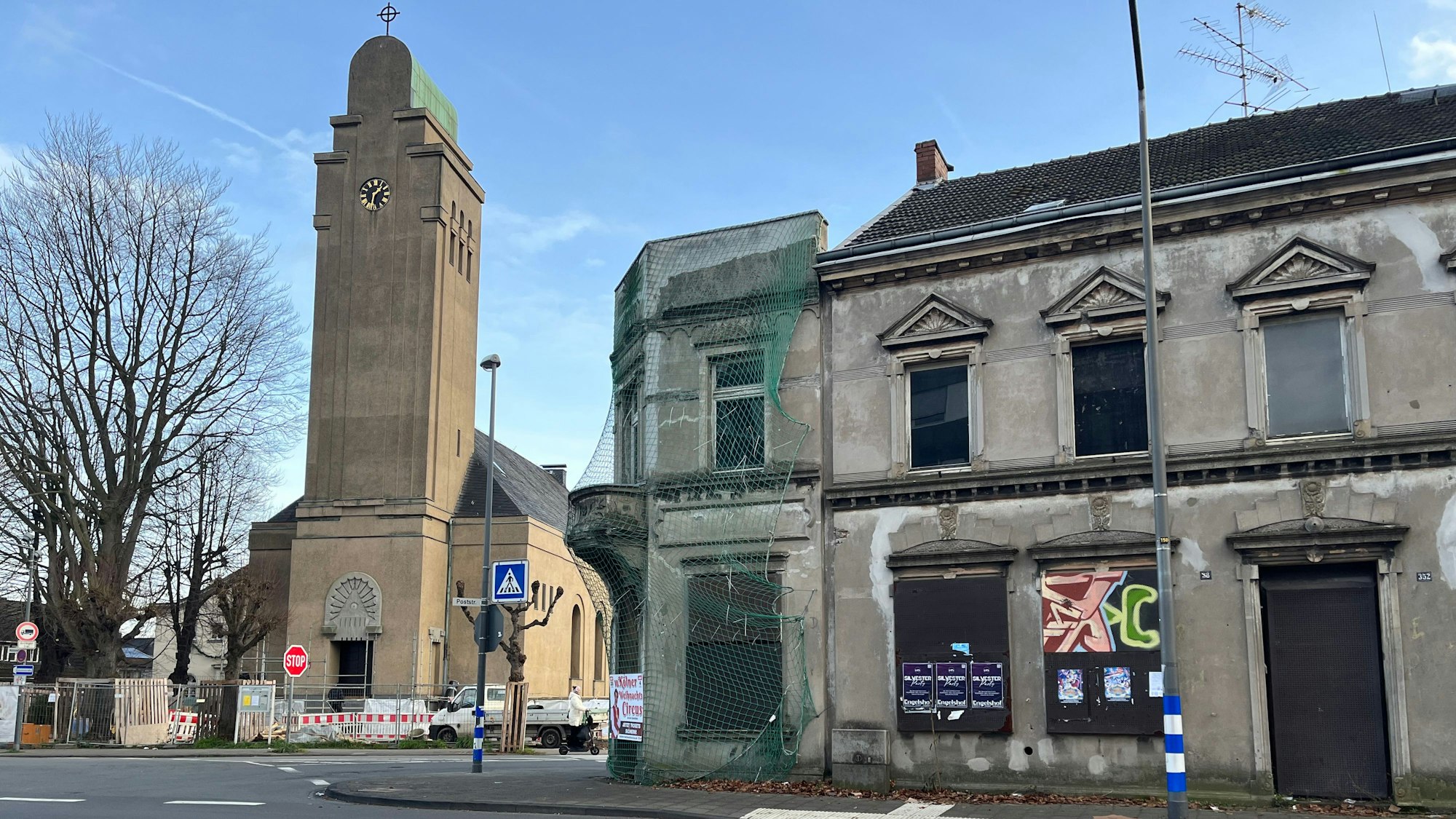 An einer Straße stehen eine Kirche und ein marodes dreistöckiges Haus, das teils von einem Schutznetz umgeben ist.