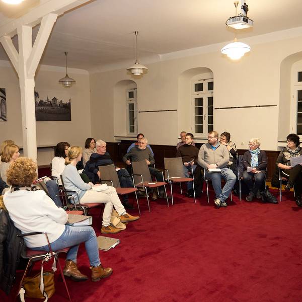 Einige Männer und Frauen sitzen im Halbkreis auf Stühlen, rechts steht der Steinfelder Pfarrer Wieslaw Kaczor.