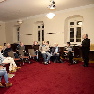 Einige Männer und Frauen sitzen im Halbkreis auf Stühlen, rechts steht der Steinfelder Pfarrer Wieslaw Kaczor.