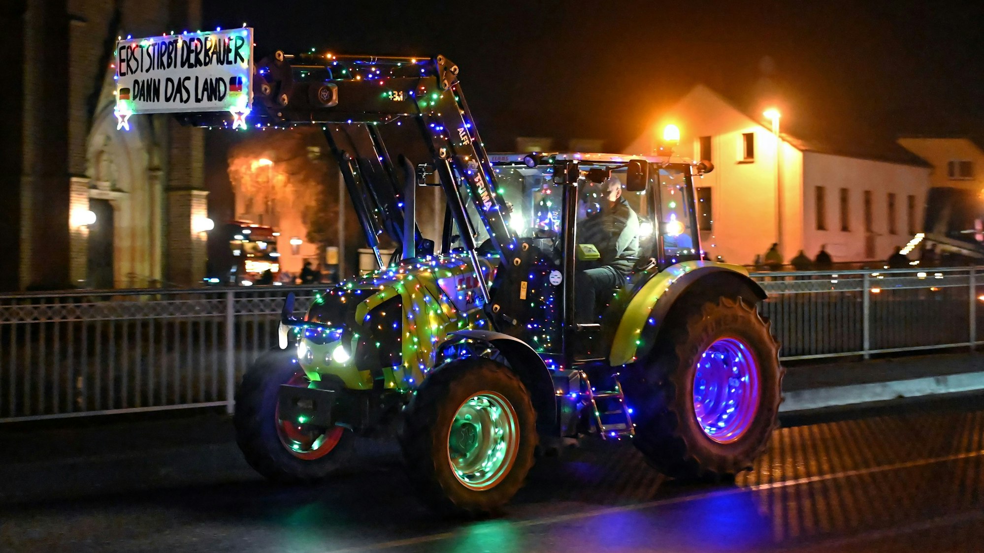 Ein Teilnehmer hat seinen Traktor mit buten Lichterketten geschmückt. An der Schaufel hängt ein Protestschild mit den Worten „Erst stirbt der Bauer, dann das Land“.