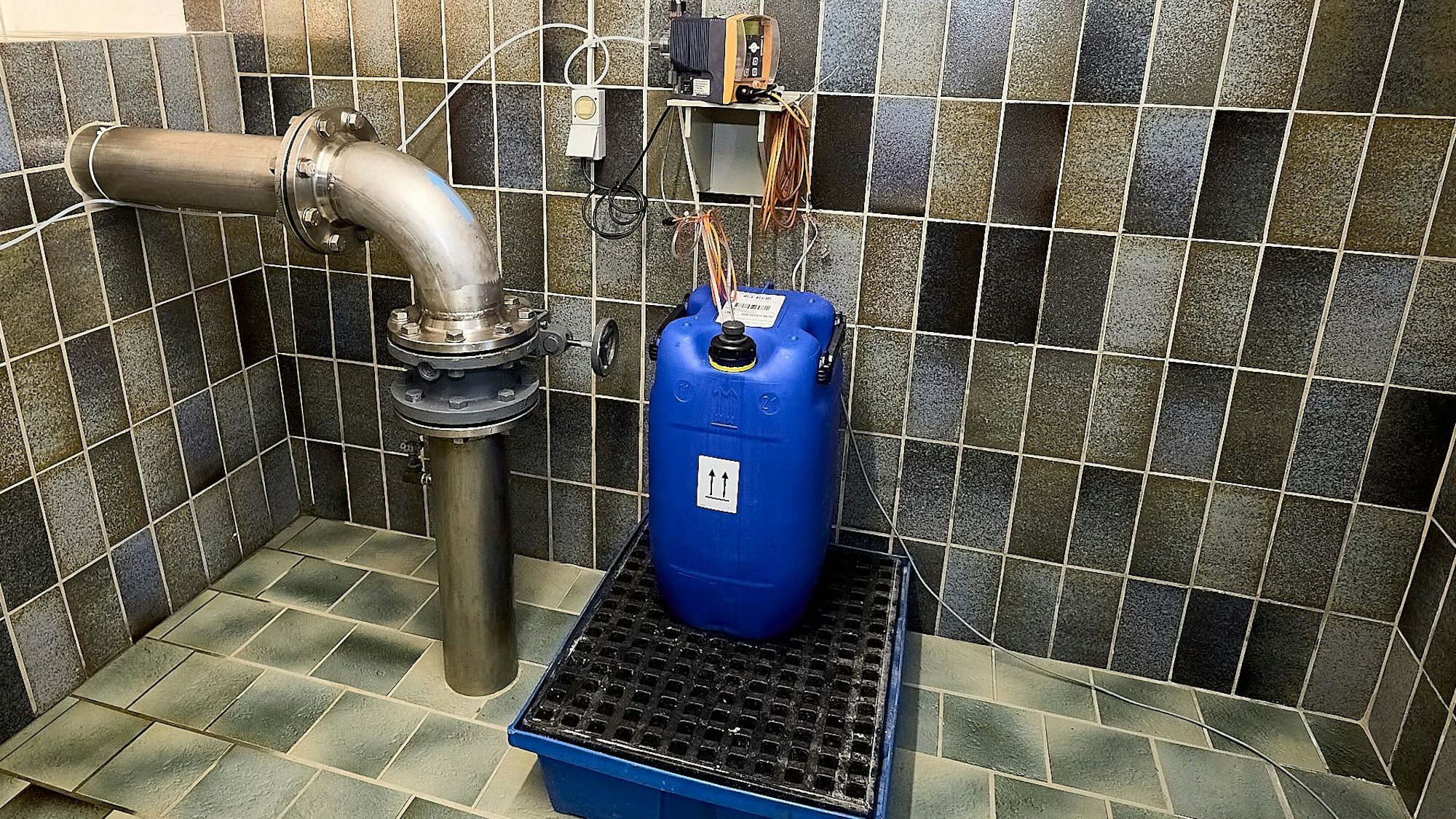 In einem grau-braun gekachelten Raum steht ein blauer Kanister, von dem aus Schläuche in ein Metallrohr führen.