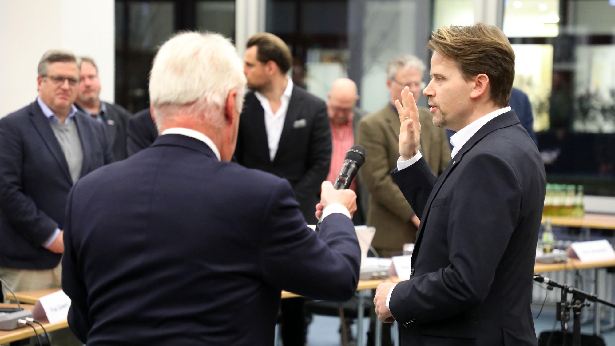 Ein Mann, dem ein Mikrofon an den Mund gehalten wird, hebt die Hand und spricht.