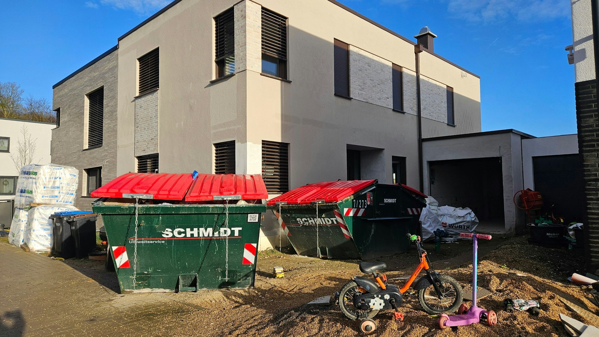 Vor einem Neubau stehen Container, auf einem Sandhaufen sind ein Kinderfahrrad und ein Kinderroller abgestellt.