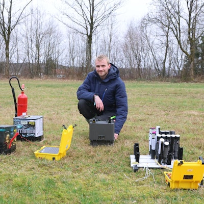 Ein junger Mann kniet neben Feuerwerkskörpern auf einer Wiese.