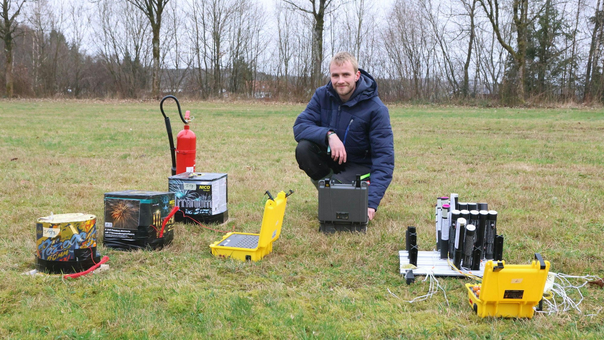 Ein junger Mann kniet neben Feuerwerkskörpern auf einer Wiese.