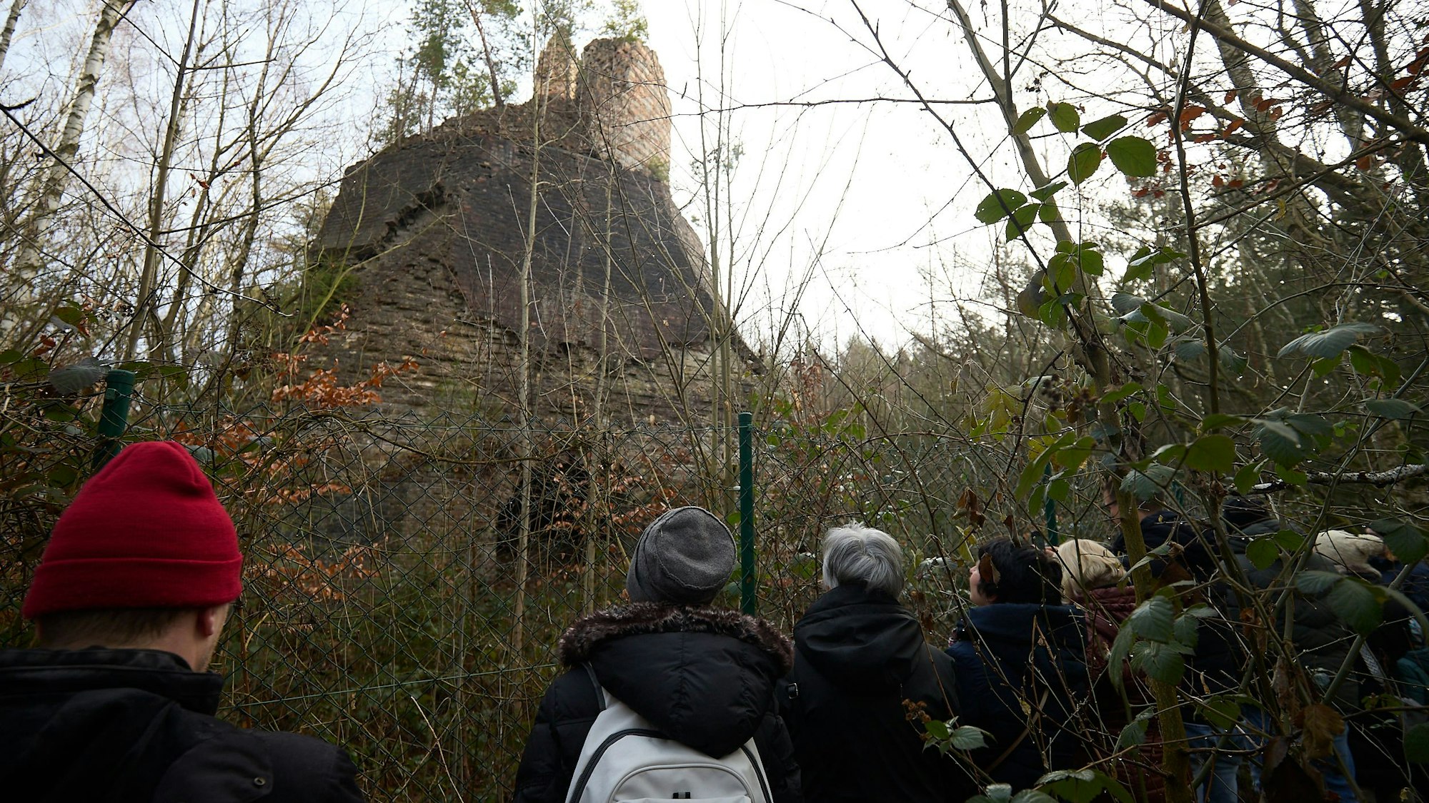 Vor dem Stumpf des Bergwerksschornsteins stehen Menschen. Sie wenden dem Fotografen den Rücken zu,