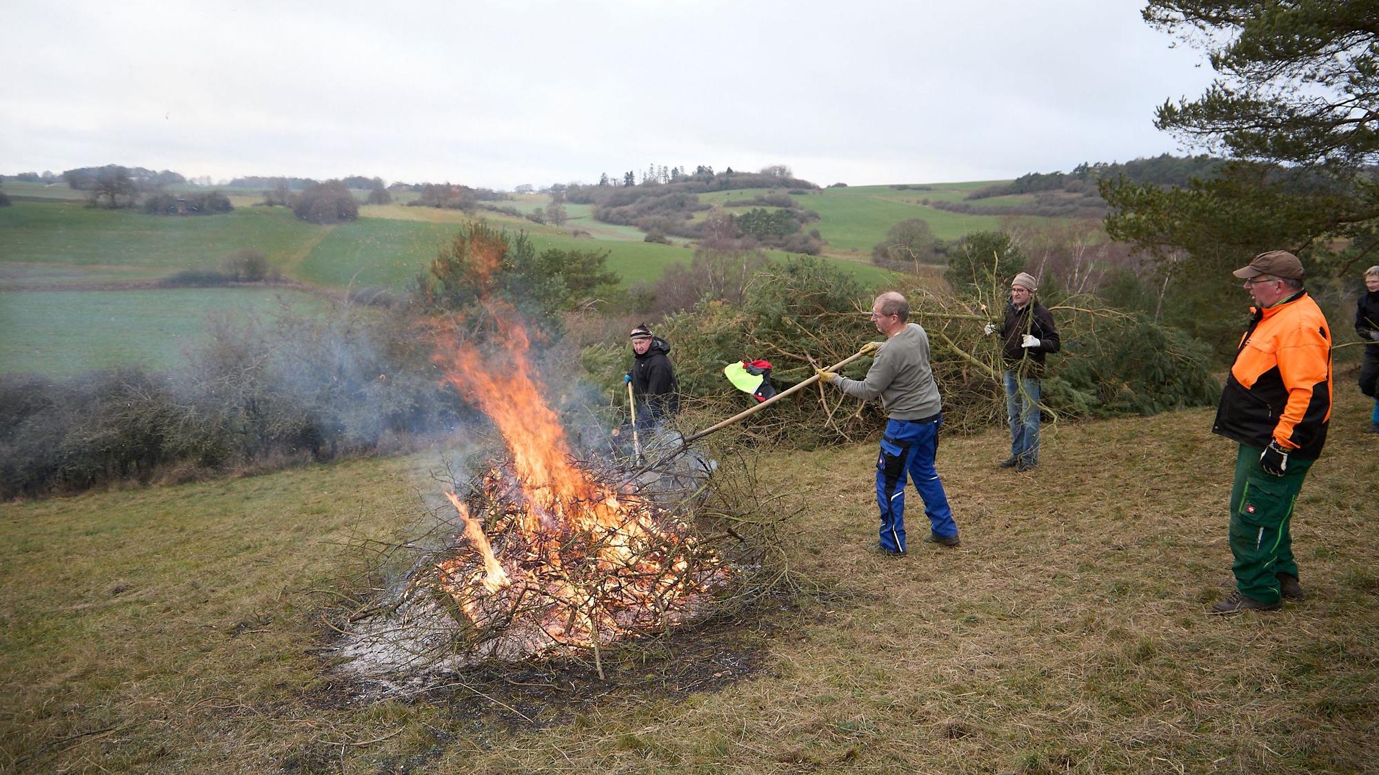 Auf einem Hügel brennt ein Feuer, Helfer bringen weiteres Strauchwerk, das verbannt werden soll.
