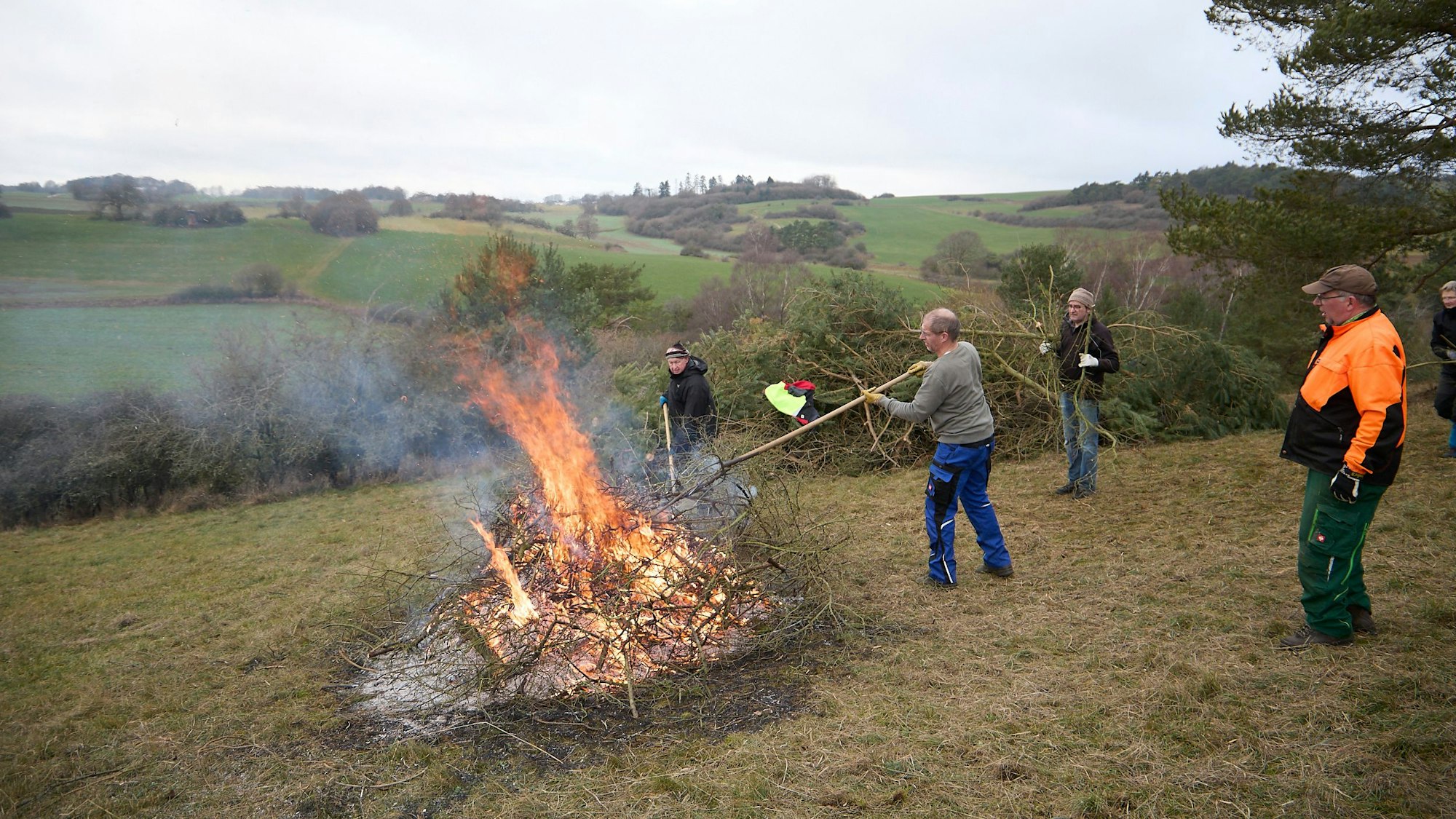 Auf einem Hügel brennt ein Feuer, Helfer bringen weiteres Strauchwerk, das verbannt werden soll.