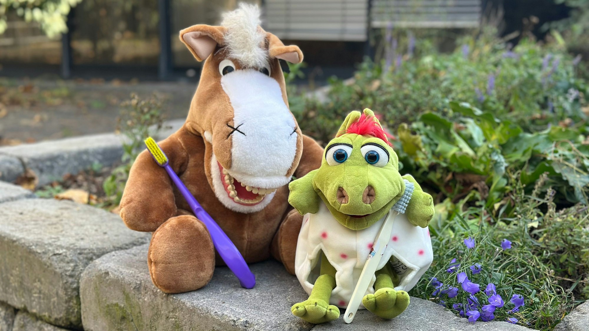 Ein Plüschpferd und eine Plüschdrache sitzend auf einer Mauer, beide halten Zahnbürsten.