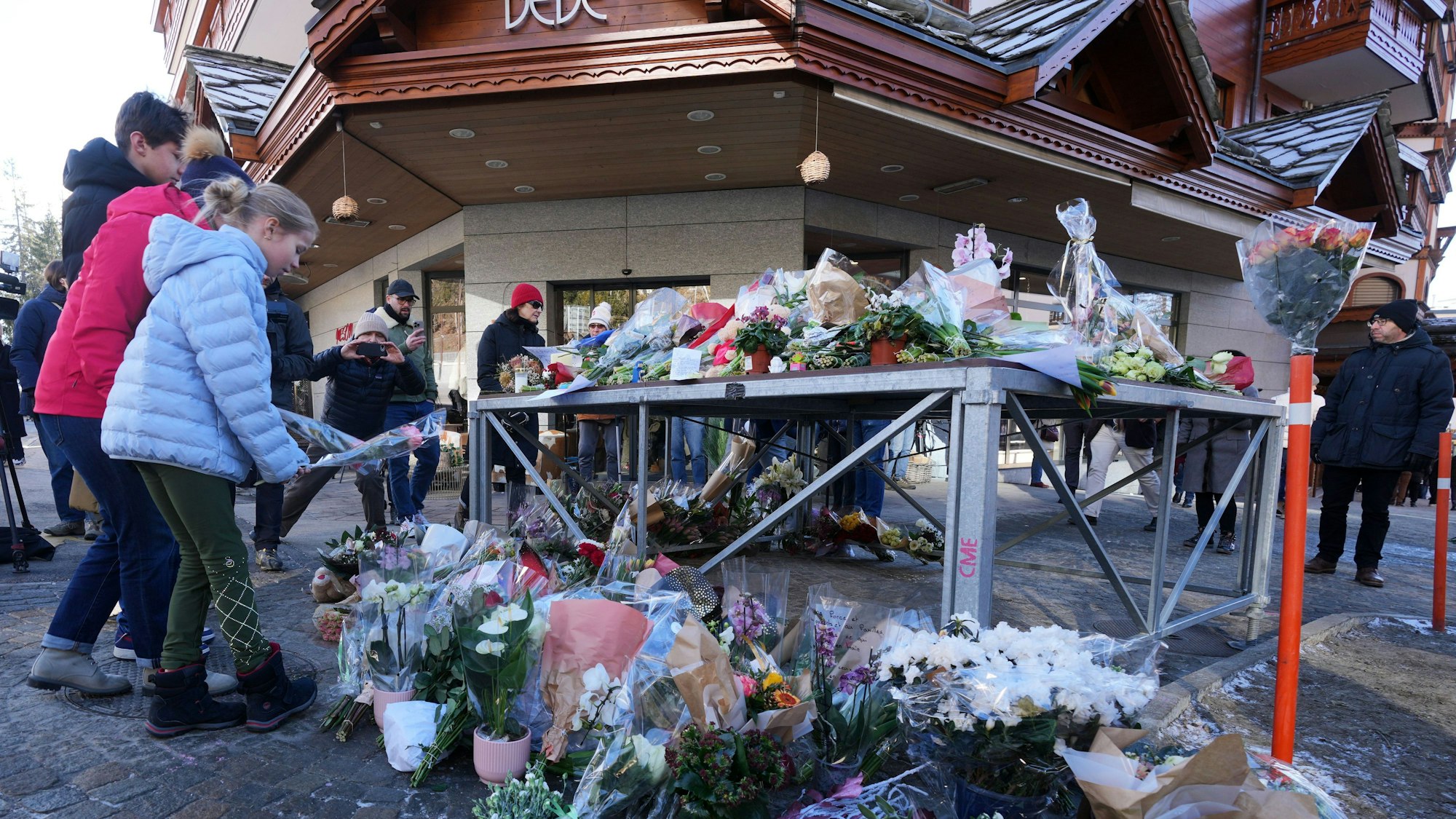 Das Bild zeigt Menschen, die Blumen und Briefe in die Nähe der abgeriegelten Bar Le Constellation, wo ein verheerendes Feuer während der Silvesterfeierlichkeiten in Crans-Montana in den Schweizer Alpen Tote und Verletzte forderte, ablegen. Foto: Antonio Calanni/AP/dpa