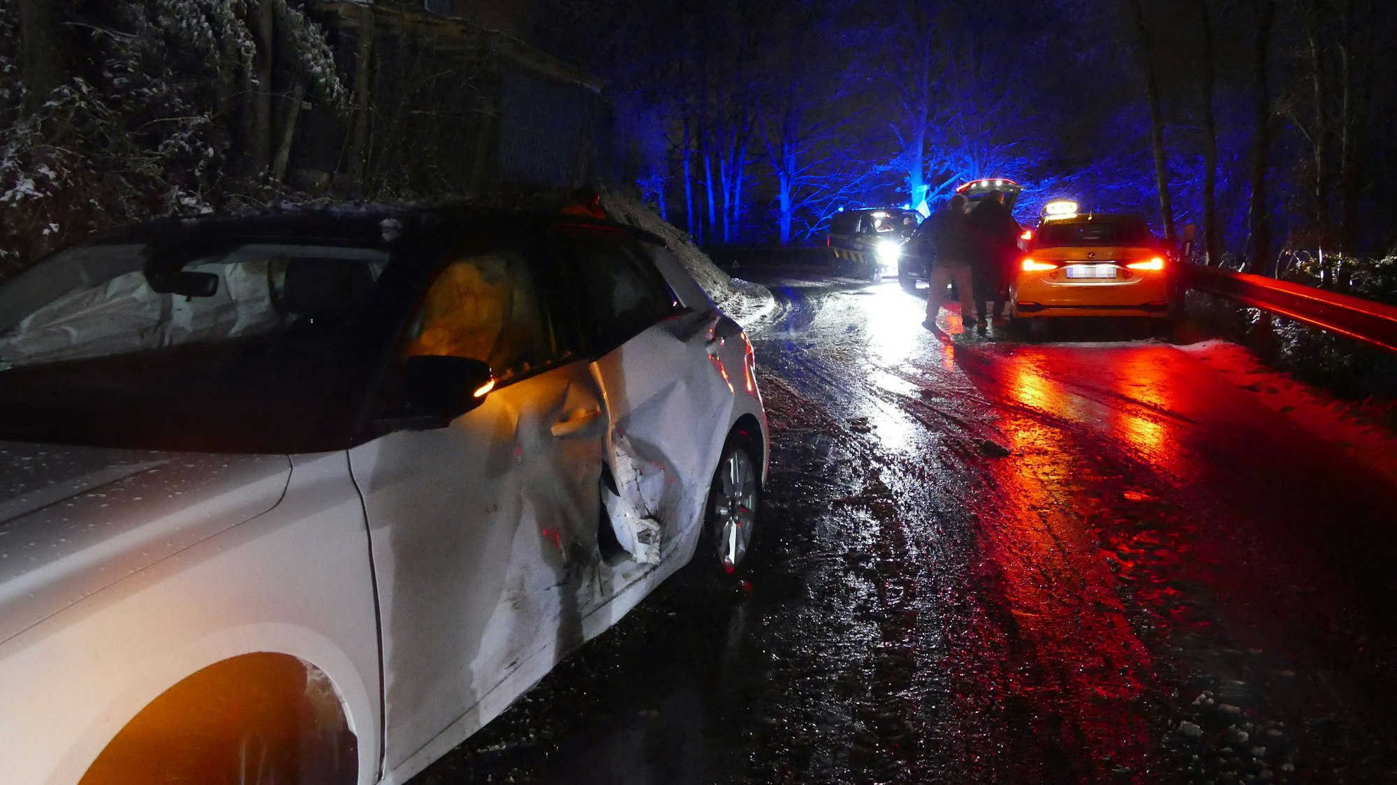 Ein beschädigtes Auto an einer Unfallstelle. Es ist dunkel, die Straße ist nass.