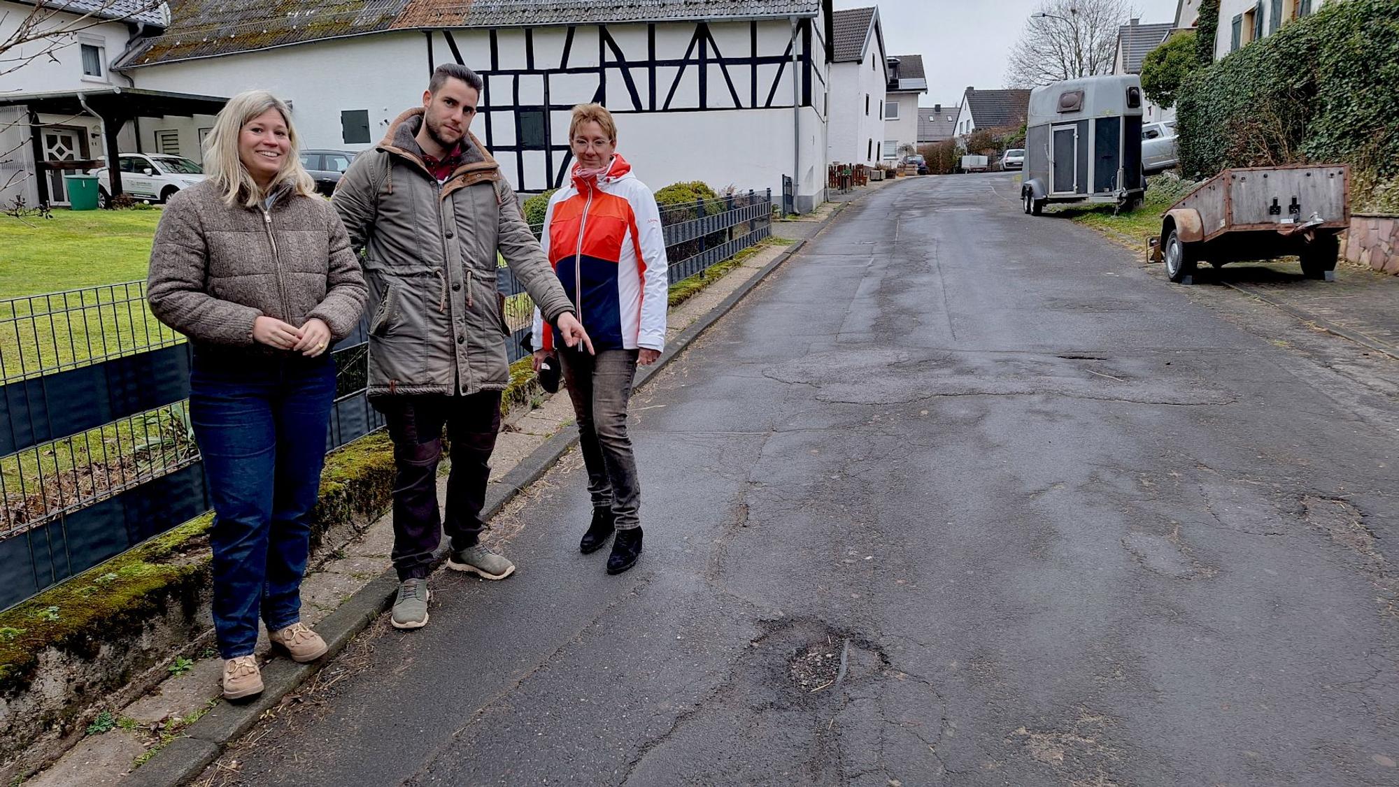 Dirk Dombrofski, der mit den beiden Frauen am Straßenrand steht, zeigt auf ein Schlagloch.
