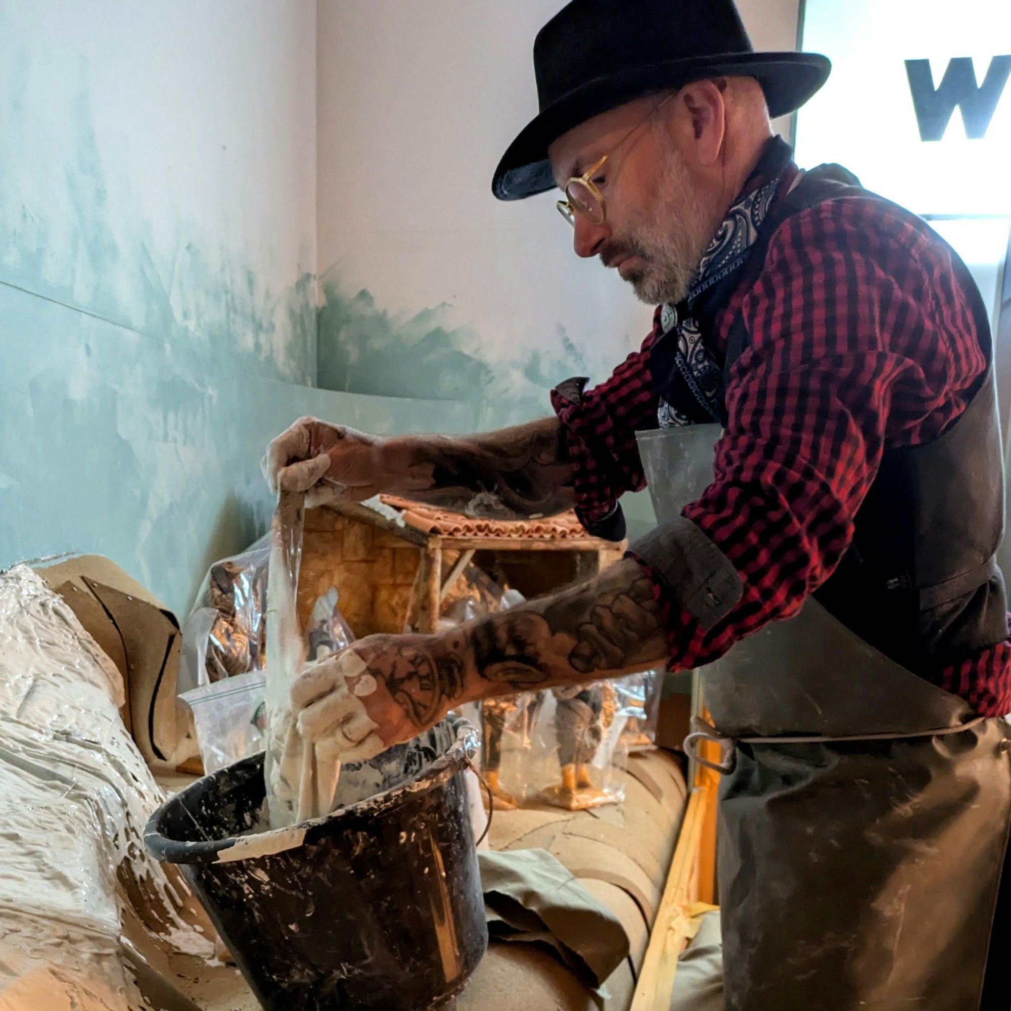 Olivier Jaubert trägt eine Schürze über einem karierten Hemd. In einer Werkstatt taucht er Textilien in einen Eimer mit einer Gipsmasse, um daraus später Landschaften für Krippen zu modellieren.