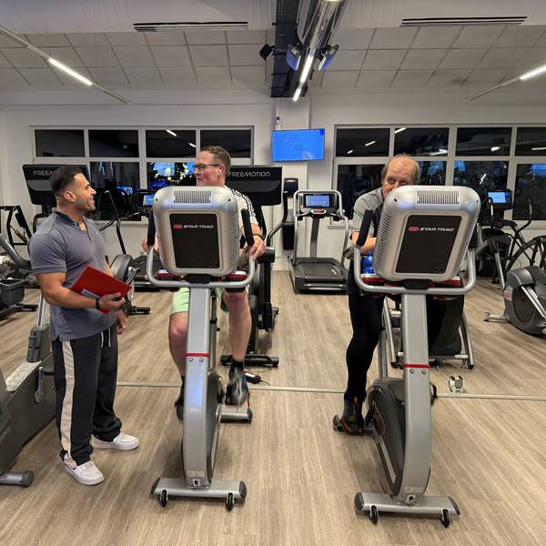 Zwei Männer sitzen im Fitnessstudio auf dem Fahrrad, während der Trainer daneben steht.