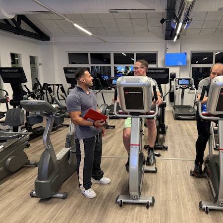 Zwei Männer sitzen im Fitnessstudio auf dem Fahrrad, während der Trainer daneben steht.