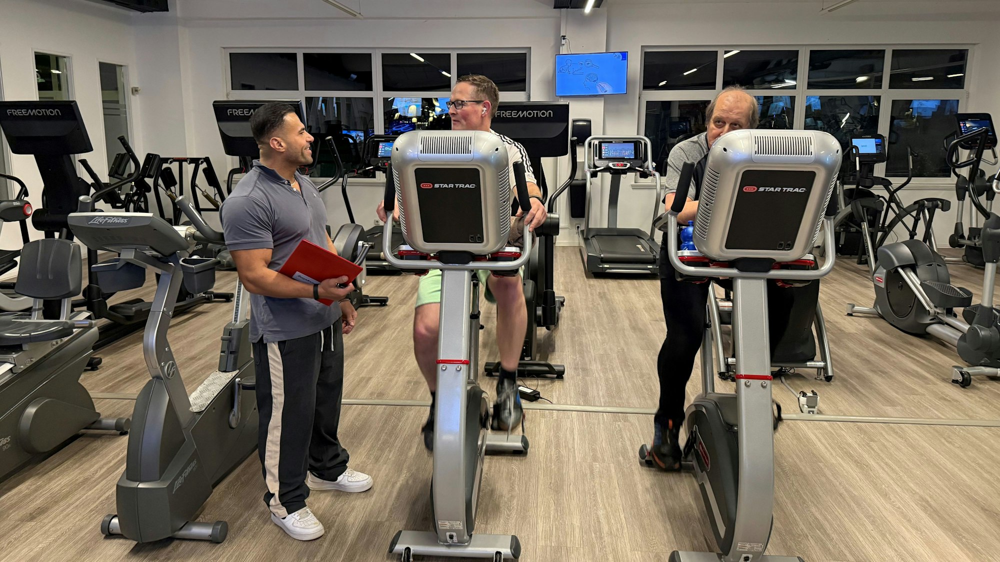 Zwei Männer sitzen im Fitnessstudio auf dem Fahrrad, während der Trainer daneben steht.