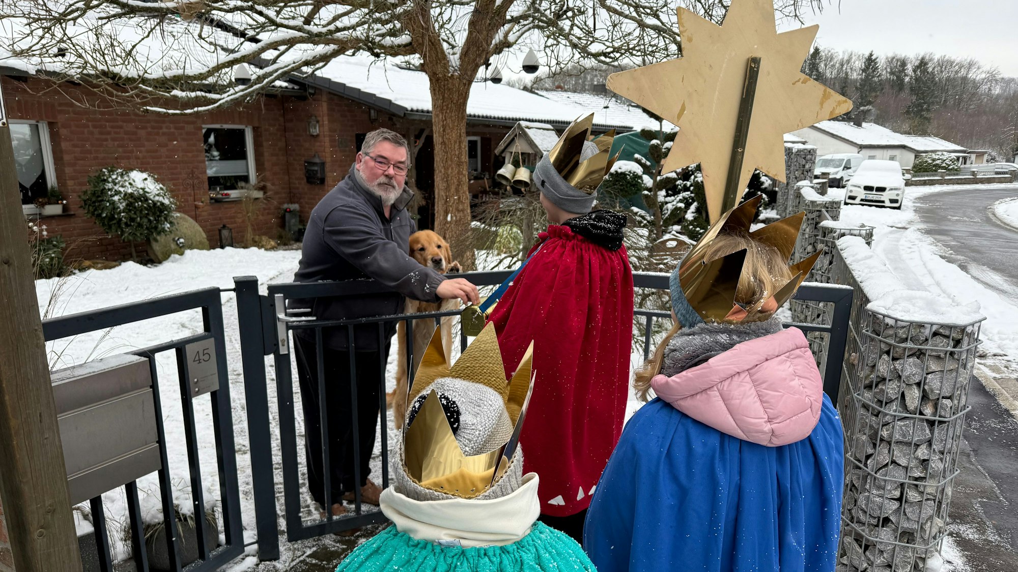 Drei Sternsinger stehen vor einem Gartenzaun und nehmen eine Spende von einem Mann entgegen.