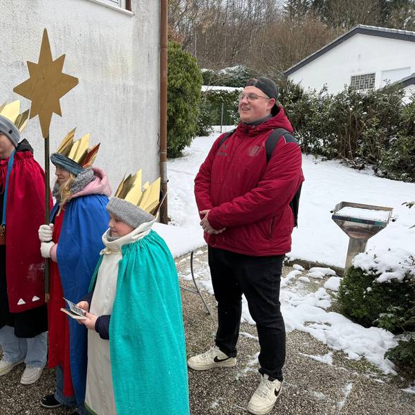 Drei Kinder stehen als Sternsinger vor einer Tür. Dahin steht ihr Begleiter